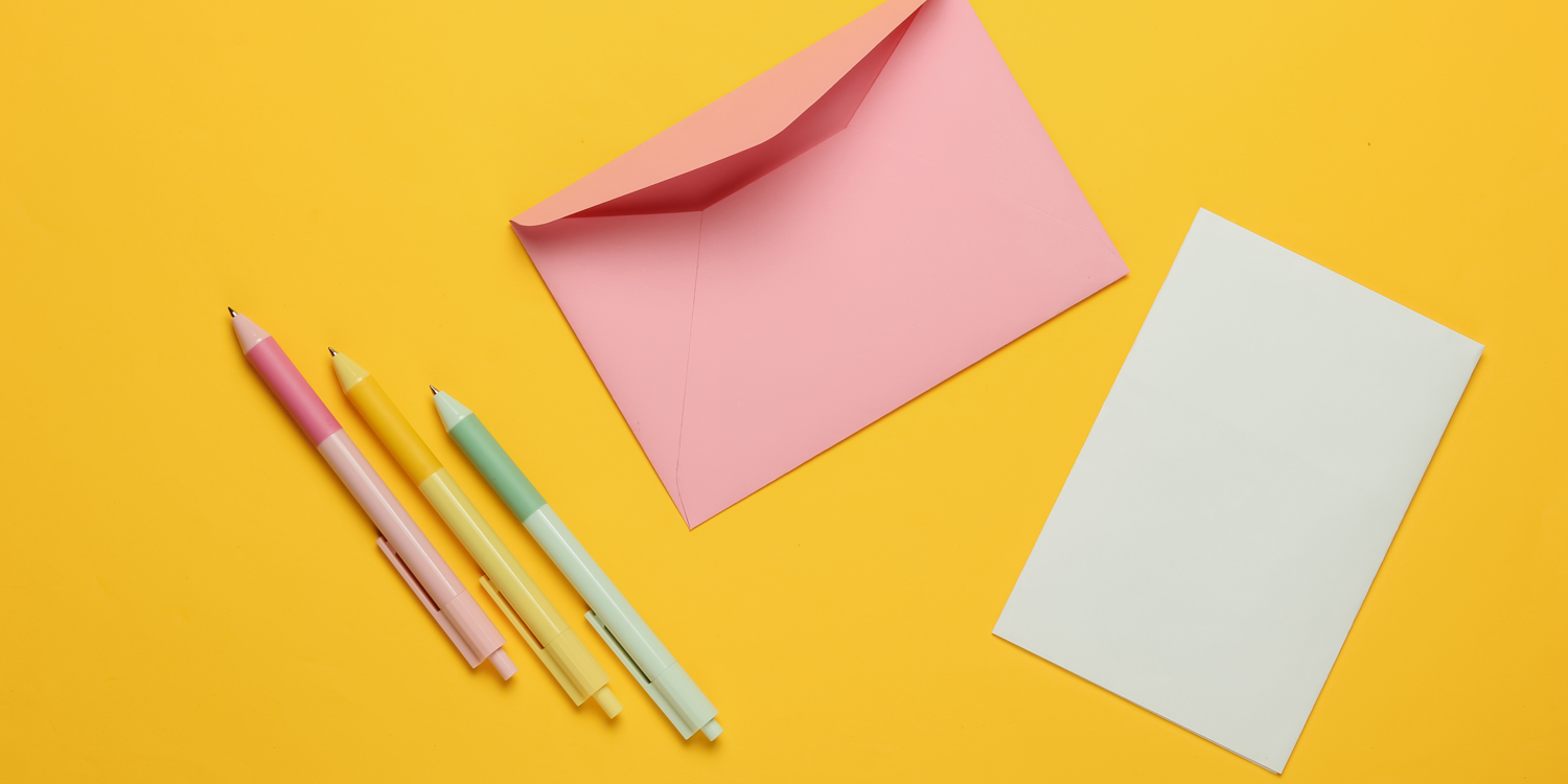 Man sieht ein rosarotes Kuvert , ein weißes Briefpapier und drei Kugelschreiber